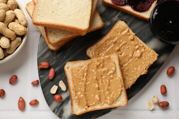 Delicious toasts with peanut butter, nuts and jam on white tiled table, top view