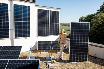 Portrait of a man standing with solar panel on a rooftop of his house during installation process, wide angle view. Owner of property installing solar panels for self consumption