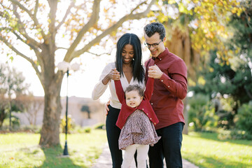 Smiling parents raising little girl by hands while standing in park