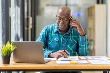 Portrait of Senior African man sitting on laptop at the home office and having a phone conversation...