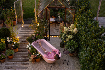 Cozy backyard with a woman bathing in pink bathtub outdoors at dusk. Concept of relaxation and beauty