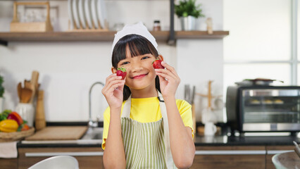 Portrait little toddler asian girl child in apron closing eyes by strawberry smiling and looking to camera. Cheerful little girl having fun holding strawberry on hands covering eyes