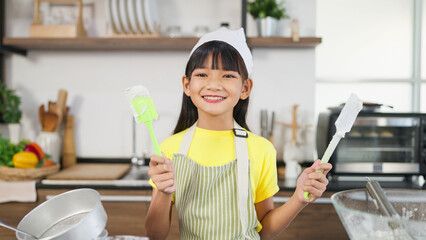 Asian little girl in apron and chef hat holding kitchen equipment on hands smiling to camera in...