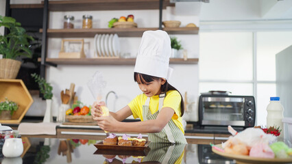 Little toddler asian girl child in apron and chef hat whipped cream decorating preparing homemade cupcakes in home kitchen. A Little girl preparing and decorating homemade cake. Children cooking - Powered by Adobe
