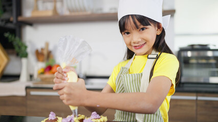 Asian little girl in apron and chef hat holding bag of whipped cream smiling to camera in kitchen at home while decorating preparing homemade cupcakes. Concept for cooking learning for kid