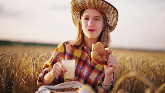 Young girl farmer in sun hat eats loaf of bread biting it drinking milk sits on wheat field with grain bag on knees. Harvesting, summer on farm, food business, agribusiness, woman worker has break.