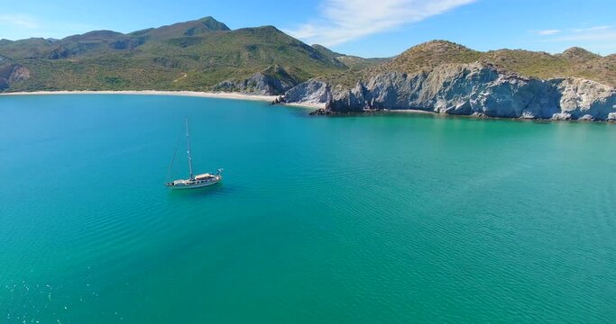 A group of monohull sailing yachts anchored in a brilliantly turquoise blue bay along a pristine beach at the base of small mountainous islands