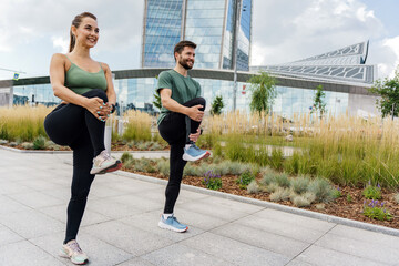 Joyful fitness duo doing leg stretches in urban park, high-rise buildings backdrop.