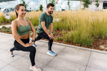 Happy couple doing lunges in city park, skyscraper reflections in background.