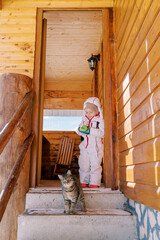 Little girl stands on the threshold of a wooden cottage and looks at a striped cat on the steps