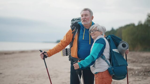 Senior hikers hike on nature autumn park holding nordic sticks talking discussing route. Tourists man woman trekking carrying backpacks with touristic equipment. Nordic walking travel tourism concept.