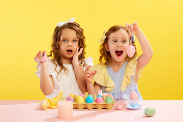 Emotional little girls, children making, decorating and painting Easter eggs against yellow background. Concept of Easter holiday, celebration, traditions, childhood, happiness