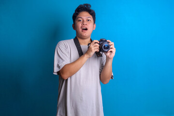 Shocked young professional photographer taking picture using digital camera on blue background