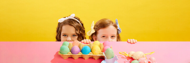 Cute little girls peaking out table and looking at colorful painted Easter eggs against yellow background. Concept of Easter holiday, celebration, traditions, childhood, happiness. Banner