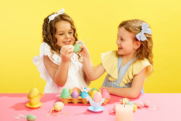 Obraz na płótnie Canvas Happy smiling little girls, kids cracking painted Easter eggs, having fun against yellow background. Concept of Easter holiday, celebration, traditions, childhood, happiness, family