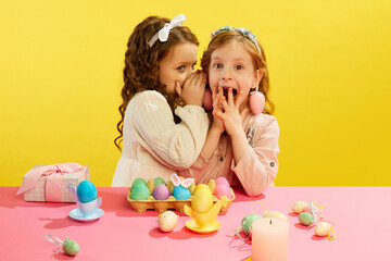 Obraz na płótnie Canvas Little girl whispering to friend on ear, kids happy and excited face against yellow background. Concept of Easter holiday, celebration, traditions, childhood, happiness