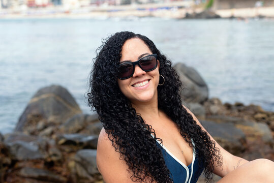 Beautiful, curly-haired woman wearing sunglasses looking at the camera against the sea in the background.