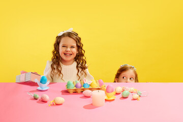 Happy little girl holding present, kid peeking out table with painted colorful painted eggs against yellow background. Concept of Easter holiday, celebration, traditions, childhood, happiness