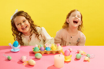 Obraz na płótnie Canvas Little girls, kids cheerfully laughing, sitting at table with decorated and painted eggs against yellow background. Concept of Easter holiday, celebration, traditions, childhood, happiness