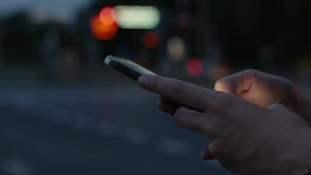 Woman uses smartphone on night street, reading social media and browsing internet. Person is holding phone in hands, pressing on display against night street with moving cars. Online addiction