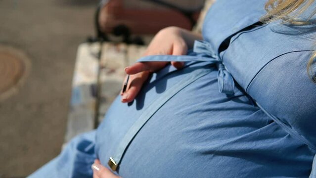 A Pregnant Woman is stroking her Belly with her Hand. A pregnant woman in a blue dress is resting on a bench. A Child pushes women in the belly.