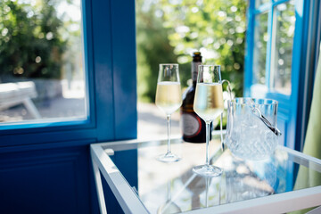 Close up of champagne glasses on transparent table. Bottle of wine and ice cooler. Blue door and...