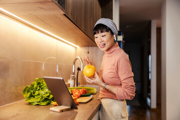 A middle-aged japanese housewife is having video call on tablet while preparing healthy meal at home.