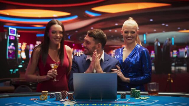Portrait of a Happy Man with Two Female Partners Betting Online on a Laptop Computer while Sitting in a Luxury  Casino. Lucky Man and Glamorous Women Celebrating Blackjack Winning Jackpot . Zoom Out