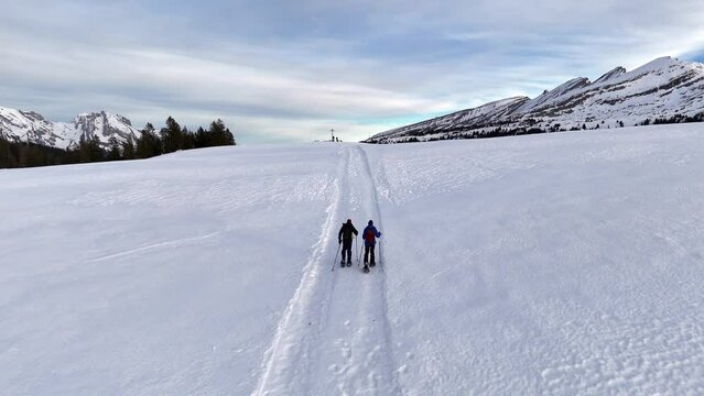 Aerial following shot of two people snowshoeing on snowy mountain with beautiful alps view in background - rising shot