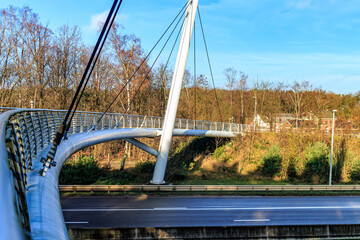 Part of cable-stayed bridge for bicycles and pedestrians over highway towards Hoge Kempen national park, bare trees against blue sky in background sunny autumn day in As Limburg, Belgium - Powered by Adobe