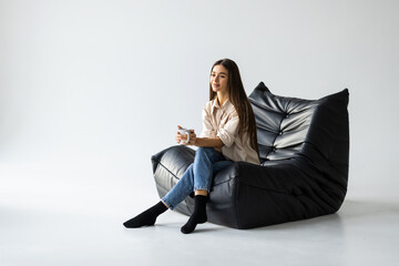 Young woman sitting on comfortable bean bag at home drinking coffee in living room.