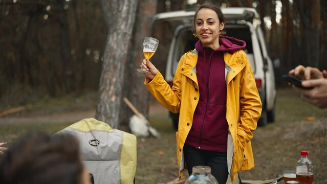 Smiling joyful woman with glass of beer relaxing outside in autumn forest with friends. Positive friends at picnic outdoors in fall woods with camper van . friends talking with beer for grilled meat.