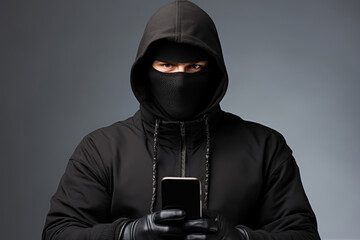 Male criminal hacker in black clothes and balaclava with phone in hand, phone scam concept - obrazy, fototapety, plakaty