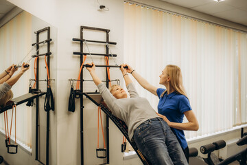 Positive senior woman doing stretching exercises on Pilates reformer as part of injury rehabilitation course. Therapeutic physical training concept - obrazy, fototapety, plakaty