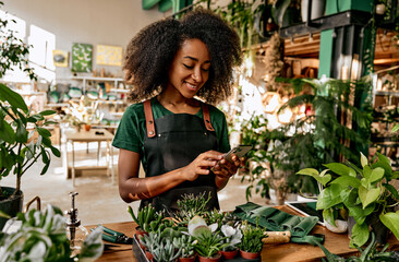 Beautiful african american woman florist in a green t-shirt and apron stands in the shop with plants on the table and scrolls the phone while working. Busy business lady taking orders online. - obrazy, fototapety, plakaty