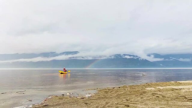 Alone man on kayak boating in Siberian Teletskoye Lake. Calm spring weather, cloudy sky and rainbow