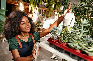 Small business. Pretty black woman with watering can in hands standing near white metal stand full...