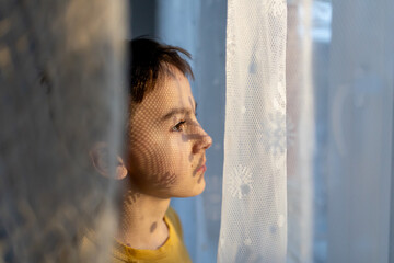 Artistic portrait of a teenage boy, standing next to a window, shadows from curtains falling on his face, making interesting shadows,