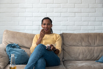 Laughing young pretty African American woman sitting on sofa, eating popcorn and watching TV at home, holding remote controller, enjoying nice movie at weekend. Business woman quality free time