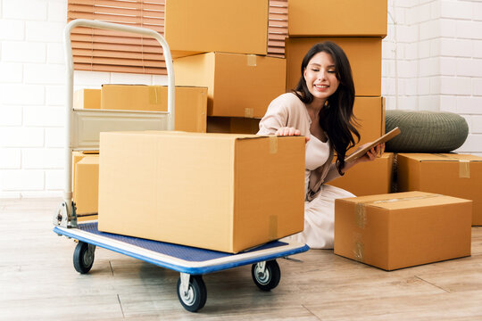 Happy Asian couple moving to a new house, a woman online shopping with a digital tablet while sitting amid cardboard boxes from the old house. Family-Moving house relocation, home moving concept.