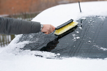 A man's hand with a brush cleaning the rear window of a car from snow in the winter season.