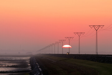 Scenic view to the Rømø causeway with 
transmission lines, fascines and red sky at sunrise. - obrazy, fototapety, plakaty