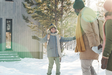 Happy cute boy in warm winterwear looking at his father while walking in front of his parents while...