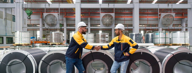 Two people shaking hands in front of large rolls of steel at an industrial facility.