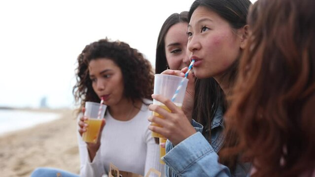 group of diverse women on the beach drinking refreshing drink having fun laughing - focus on asian woman -