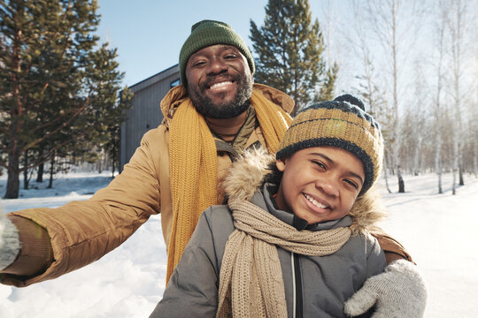Cheerful African American father and son in winterwear looking at camera in rural environment on frosty winter day while taking selfie