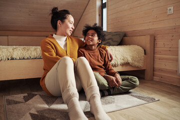 Young woman in warm casualwear sitting on the floor next to her cute son and talking to him while spending weekend or vacation in cottage - obrazy, fototapety, plakaty