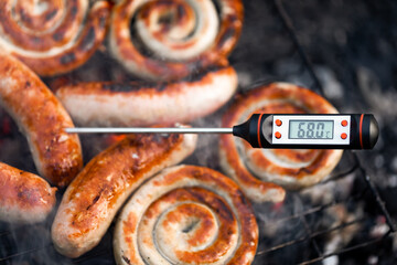 Close-up shot of a digital meat thermometer displaying a safe cooking temperature of sausages cooking on a smokey grill