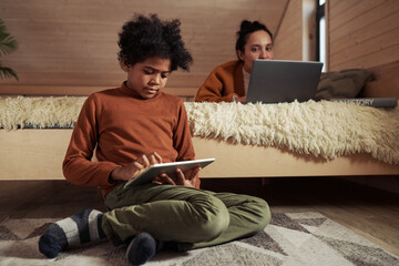 Cute serious African American boy in casualwear sitting on the floor by bed and scrolling in tablet...