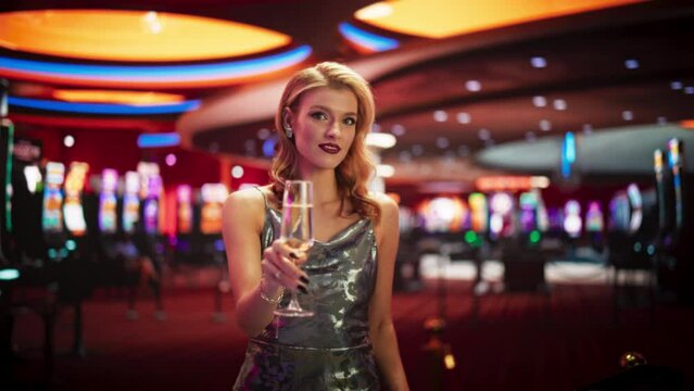 Beautiful Caucasian Woman Posing In Prestige Casino Next To Baccarat Table And Wearing Fancy Dress. Female High Stakes Player Enjoying Glass Of Champagne, Smiling, Looking At Camera in Slow Motion.